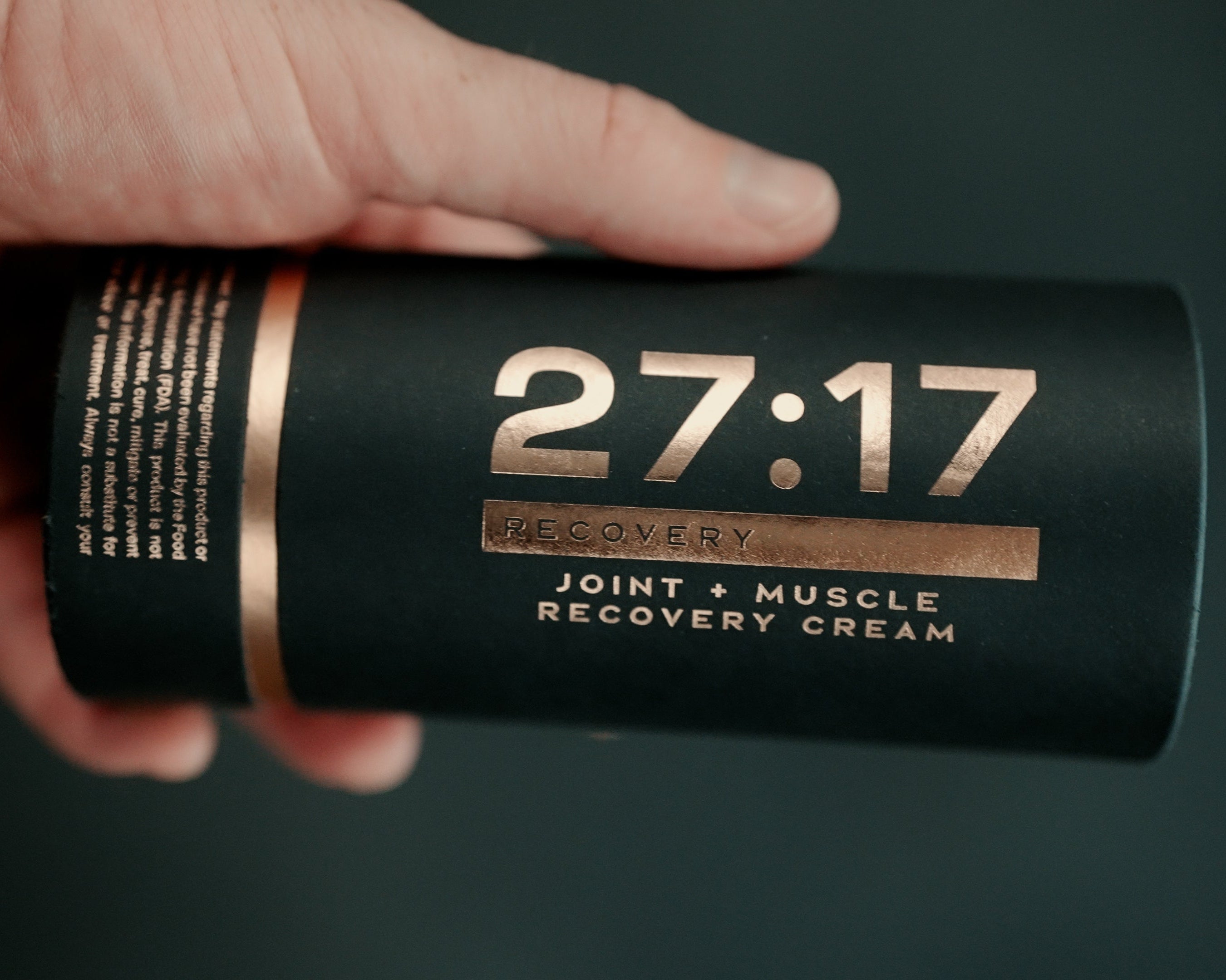 A hand holding a black cylindrical tube of recovery cream with gold text and a gold band, labeled '27:17 Recovery Joint + Muscle Recovery Cream'.