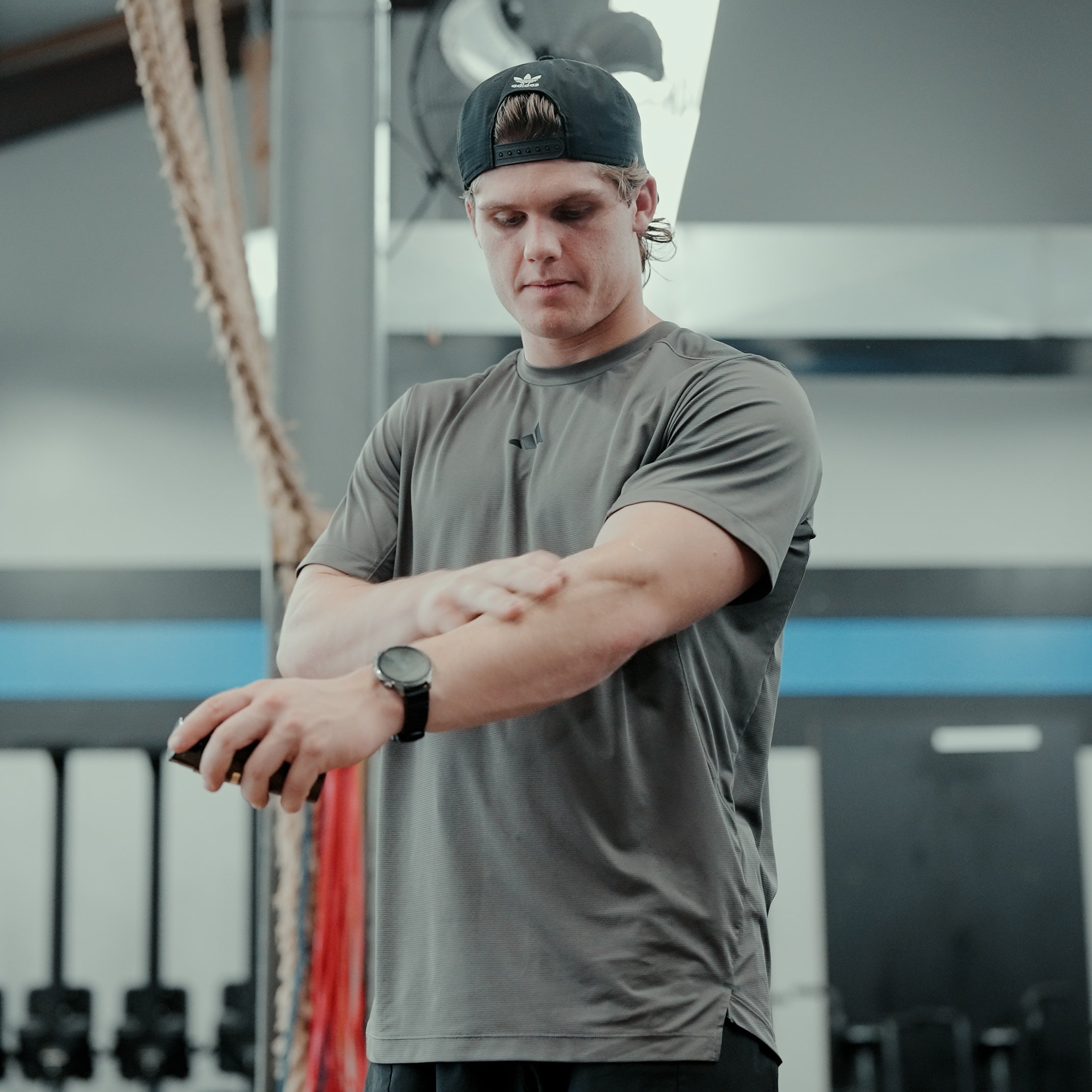 Ty Jenkins, 2026 Professional CrossFit Games Athlete in a gym wearing a gray t-shirt and a black baseball cap, looking down and applying 27:17 Recovery Joint + Muscle Cream on his forearm after a hard workout training session.
