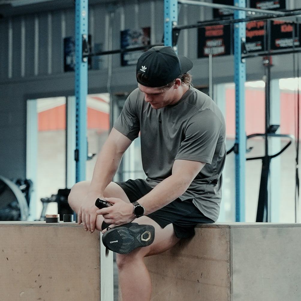Ty Jenkins, Professional CrossFit Games Athlete in a gym wearing a gray t-shirt and a black baseball cap, looking down and applying 27:17 Recovery Joint + Muscle Cream on his leg after a hard workout training session.